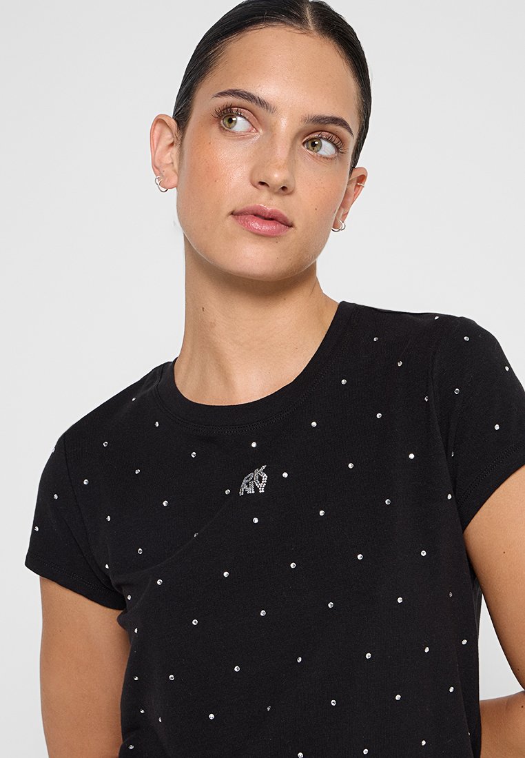 Jeune femme aux cheveux bruns portant une chemise noire à manches courtes décorée de petits points argentés et d'un petit emblème argenté près du col.