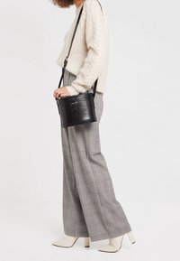 Mustankankkeinen tekonahkainen ämpärilaukku, jossa on teksturoitu pinta, pitkä säädettävä olkahihna ja hienovaraiset metallidetaljit.