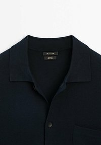 Chemise en maille bleu marine foncé avec col, boutons sur le devant et poche poitrine, avec étiquette de marque à l'intérieur du col.