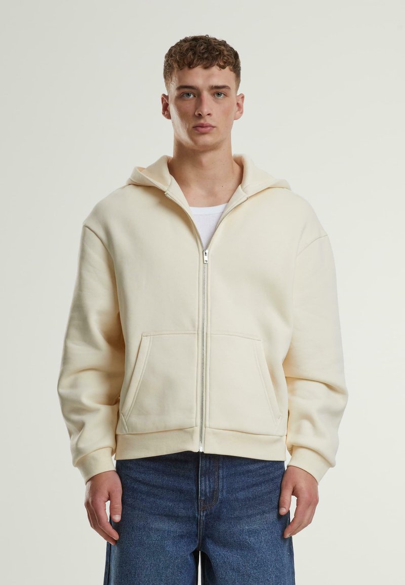 Krem fôret zip-hette med hette og foran lommer, laget av mykt stoff. Har ribbede mansjetter og nederkant. Kombinert med blå denim jeans.