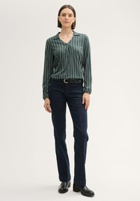 Grün gestreifte Bluse mit Kragen, kombiniert mit dunkelblauen Röhrenjeans und schwarzen Stiefeletten, vor einem neutralen Hintergrund.