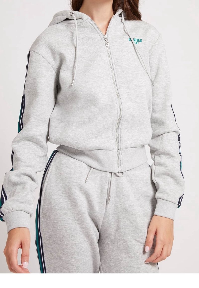 Femme portant un sweat à capuche gris clair zippé et un pantalon de survêtement assorti avec des rayures bleues et noires sur les manches et les jambes.