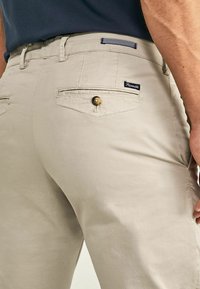 Beige cotton trousers featuring back pockets with stitched detailing and a button accent, displaying a smooth texture and casual design.