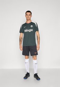 Mørkegrøn atletisk T-shirt med et hvidt og sort logo, parret med sorte shorts med en pink accent. Hvide sokker og sorte fodboldstøvler fuldender looket.