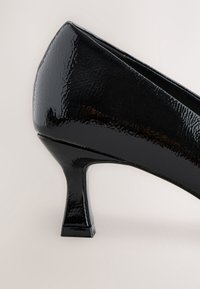 Escarpin femme en cuir verni noir avec un talon bas de forme carrée sur un fond neutre.