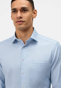 Camicia azzurro chiaro con bottoni in tessuto testurizzato, dotata di taschino sul petto, colletto classico e bottoni bianchi.