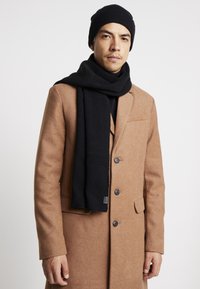 Black knit beanie and scarf, worn with a brown wool coat. Scarf features ribbed texture, coat has a tailored design with large buttons.