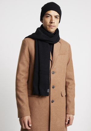 Black knit beanie and scarf, worn with a brown wool coat. Scarf features ribbed texture, coat has a tailored design with large buttons.