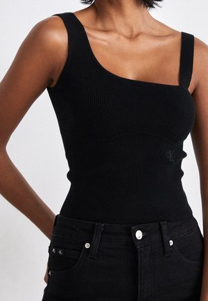 Frau trägt ein schwarzes, geripptes ärmelloses Oberteil mit asymmetrischen Trägern und schwarze, hoch taillierte Jeans vor einfarbigem Hintergrund.