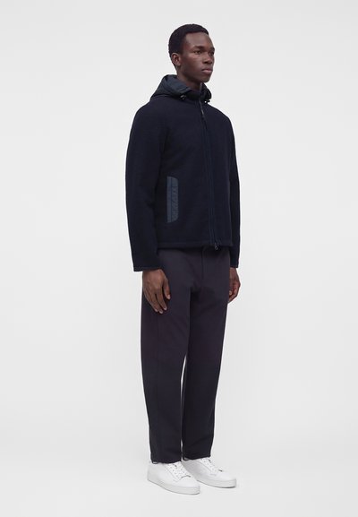 Homme debout portant une veste à capuche noire zippée, un pantalon foncé et des baskets blanches sur un fond clair uni.