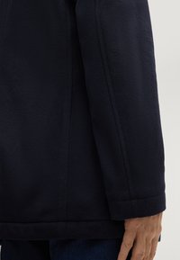 Giacca in pile blu navy con superficie testurizzata e taglio dritto. Dettagli delle cuciture visibili ai polsini e all'orlo, abbinata a jeans in denim.
