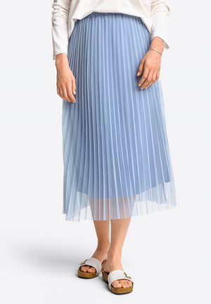 Woman wearing a light blue pleated midi skirt, white long-sleeve top, white slide sandals with gold buckle, standing on a white background.