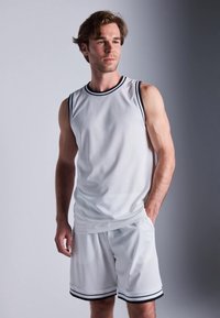 Ung man som står i vit ärmlös baskettröja och matchande shorts med svarta ränder, ser lätt åt sidan.