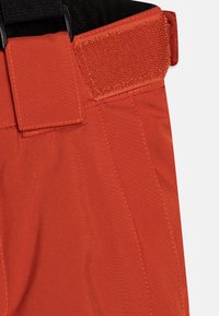 Orange, strukturerad tyg med en slät finish. Har ett kardborreband, förstärkt sömnad och en ren, definierad skärning vid midjan.