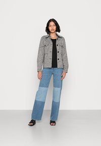 Casaco cinza texturizado com botões pretos e bolsos no peito, combinado com jeans em retalhos azul claro e sandálias de salto aberto pretas.