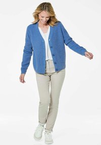 Blaues Strickcardigan mit V-Ausschnitt und Knopfschluss, kombiniert mit beigen, figurbetonten Hosen und weißen Sneakers. Einfaches, strukturiertes Stoffdesign.