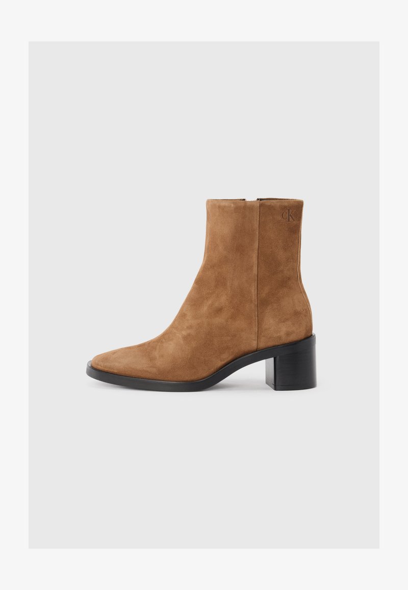 Stivale alla caviglia in suede marrone, con punta affusolata, tacco medio a blocco e discreto dettaglio del logo impresso. Chiusura con zip lungo il lato.