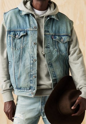 Homme portant un sweat à capuche gris clair et un gilet en denim bleu, tenant un chapeau à large bord marron foncé sur un fond beige.