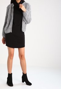 Giubbotto bomber grigio in maglia con tessuto strutturato, con cerniere, indossato su un vestito aderente nero a collo alto. Stivali neri alla caviglia con tacco a blocco completano l'outfit.