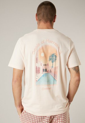Homme aux cheveux courts portant un T-shirt beige clair montrant une illustration pastel d'une piscine, d'un palmier et d'arcs intitulée « Oasis de Marrakech ».