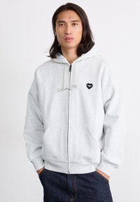 Felpa grigia con zip realizzata in cotone. Presenta una tasca frontale, cappuccio con cordino e un piccolo logo a forma di cuore nero sul petto sinistro.