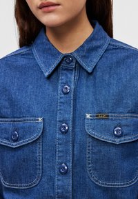 Sötétkék farmering gombos elülső résszel, két mellzsebbel és kontrasztos varrással. A zsebén márkajelzett címke található.