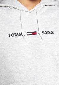 Lichtgrijze hoodie met een voorzak, voorzien van "TOMMY JEANS" in zwarte borduuring met rode en witte accenten op een horizontale band.