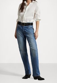 Nainen, joka käyttää valkoista napitettua paitaa, joka on tungettu sinisiin suoriin farkkuihin, mustalla vyöllä ja mustilla korkokengillä varustetuilla nilkkasaappailla.