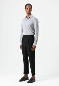 Grijs overhemd met lange mouwen en knopen, zwarte geplooide broek en zwarte loafers met strikdetail, gepresenteerd tegen een witte achtergrond.