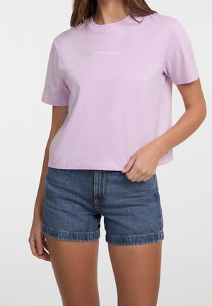 Kobieta w krótkim lawendowym t-shircie z logo "GUESS JEANS" i wysokim stanem niebieskich szortów dżinsowych stoi na jednolitym tle.
