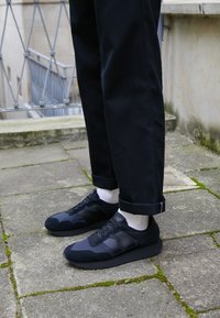 Černé tenisky se semišovými a síťovinovými panely. Zaoblená špička a šněrovací design. Nosené s černými kalhotami, které vystavují ohrnutý lem a bílé ponožky.