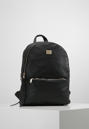 Sac à dos en cuir noir avec poche avant zippée, tirettes de fermeture éclair dorées, petite plaque logo dorée et poignée supérieure, exposé sur un socle blanc.