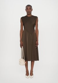 Rochie crem fără mâneci cu un detaliu geometric, dintr-o țesătură fluidă, și cu o lungime până la genunchi. Asortată cu o geantă mică din material împletit bej și cu toc.