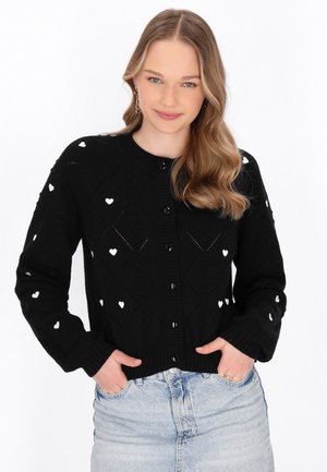 Musta neulecardigan, jossa on teksturoitu timanttikuvio ja valkoisia sydänmotiiveja. Siinä on pyöreä pääntie, nappikiinnitys ja resorit.