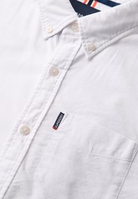 Witte blouse met een klassieke kraag, voorzien van een enkel borstzakje en een "Superdry" label met rode en blauwe accenten.