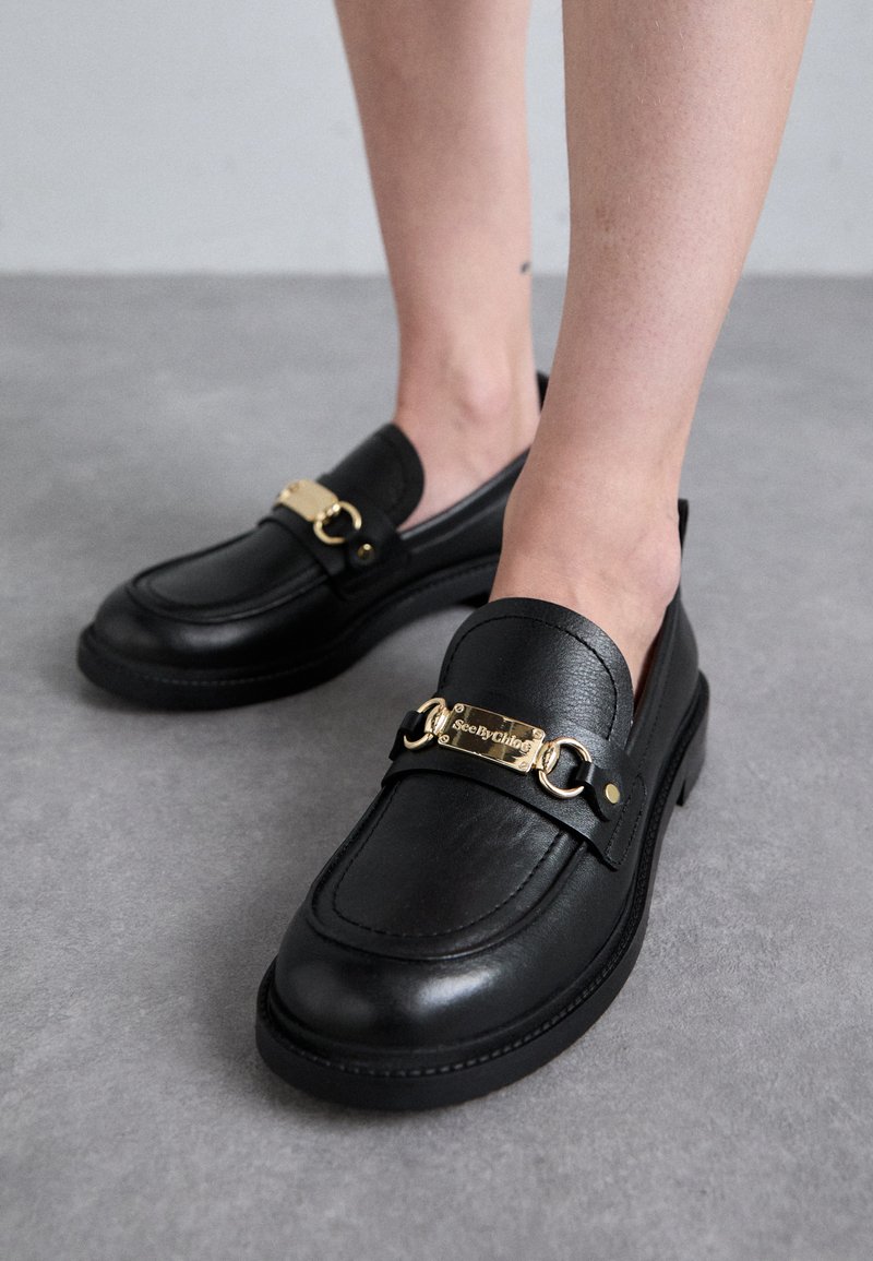 Mustad nahast loafers kuldsete detailidega paljajalu hallil põrandal.