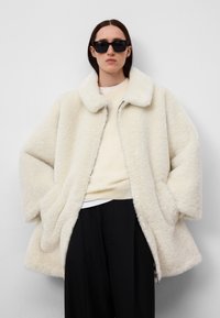 Crème-kleurige schapenvachtjas met een zachte textuur, brede kraag en ritssluiting. Model draagt een lichte trui en zwarte broek, met een zonnebril.