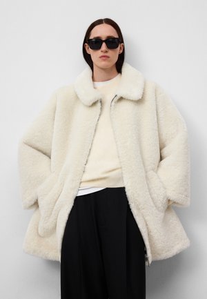 Cappotto in shearling color crema con una texture morbida, colletto ampio e chiusura con zip. Il modello indossa un maglione chiaro e pantaloni neri, con occhiali da sole.