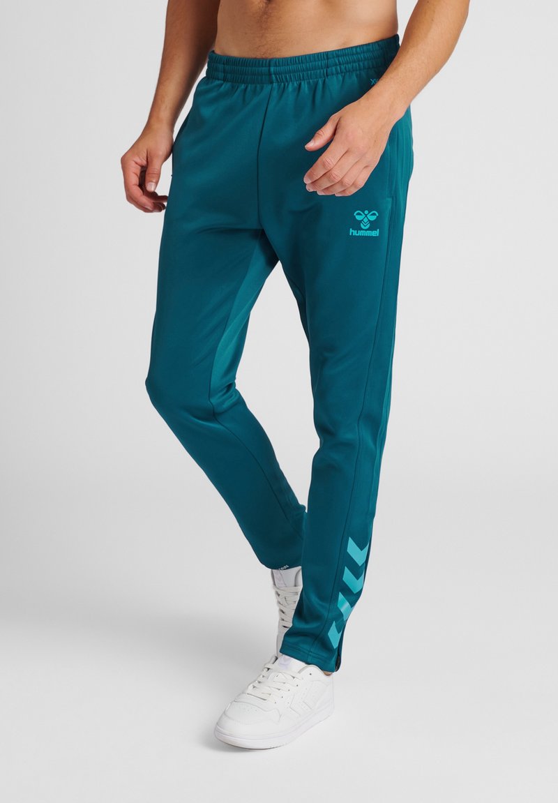 Teal sportbroek met een trekkoord in de taille, zijstrepen en een logo aan de voorkant, gemaakt van lichtgewicht, ademend materiaal. Witte schoenen worden gedragen.