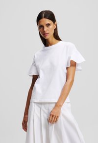 Tricou alb din bumbac, cu mâneci scurte și volănașe, și decolteu rotund. Prezentă o broderie subtilă și o croială lejeră. Asortat cu pantaloni albi.