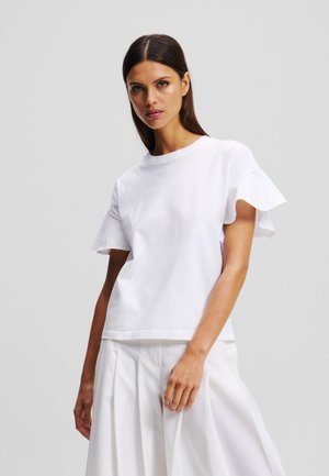 Weißes Baumwoll-T-Shirt mit kurzen, gerüschten Ärmeln und rundem Halsausschnitt. Verfügt über dezente Stickerei und einen lockeren Schnitt. Kombiniert mit weißen Hosen.