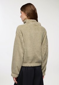 Kurtka z sztucznego futra w oliwkowym kolorze, o skróconym kroju, z wysokim kołnierzem i opadającymi ramionami. Teksturowana powierzchnia z widocznymi szwami i mankietami.