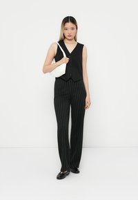 Gilet noir ajusté avec un décolleté en V et des boutons, associé à un pantalon noir à rayures. Le modèle tient un petit sac à main blanc. Chaussures plates noires.