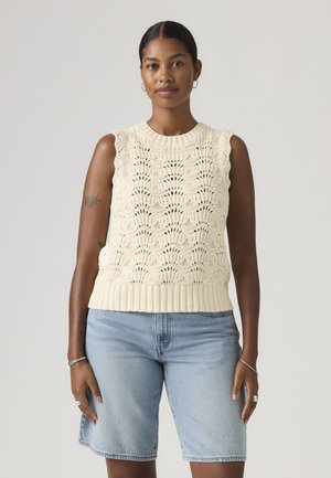 Donna con i capelli raccolti che indossa un top lavorato a maglia senza maniche color crema e pantaloncini di jeans azzurri, in piedi contro uno sfondo chiaro e semplice.