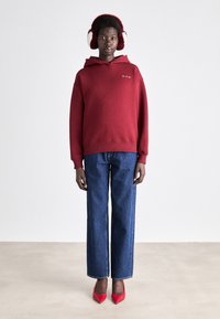 Bordeauxfarbene Kapuzen sweatshirt mit lockerer Passform, kombiniert mit geraden, dunklen Jeans und roten, spitzen Pumps. Flauschige Ohrenwärmer werden getragen.