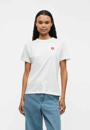 White cotton t-shirt with short sleeves and round neckline, featuring a small red heart logo on the left chest. Worn with light blue jeans.