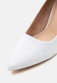 Κοντινό πλάνο λευκού γυναικείου παπουτσιού με μύτη σε σχήμα αιχμής, από γκλίτερ, με μπεζ εσωτερική επένδυση σε ανοιχτό φόντο.