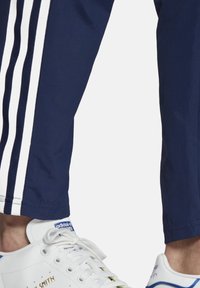 Marineblaue Jogginghose mit weißen Seitenstreifen, taillierter Schnitt; getragen mit weißen Turnschuhen mit blauen Akzenten und strukturierten Schnürsenkeln.