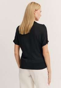 Schwarzes kurzärmliges Strickoberteil mit geripptem Saum, glatter Textur und lockerer Passform, von hinten gesehen mit glattem Haar und dezenten Ohrringen.