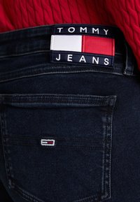 Jeans de mezclilla azul oscuro con una etiqueta de Tommy Jeans y un pequeño logo en el bolsillo trasero, combinados con una blusa de punto roja.